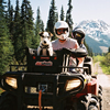 A man and his dog sit and pose for the camera on an ATV on a trail through the forest and mountains