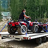 People driving trucks loaded with ATVs near a woodland area