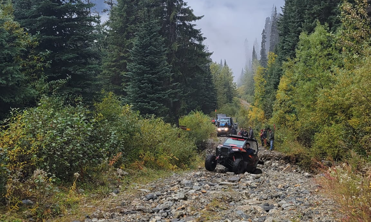 Side-by-side ATVs descend a rocky trail with tall green trees on both sides.