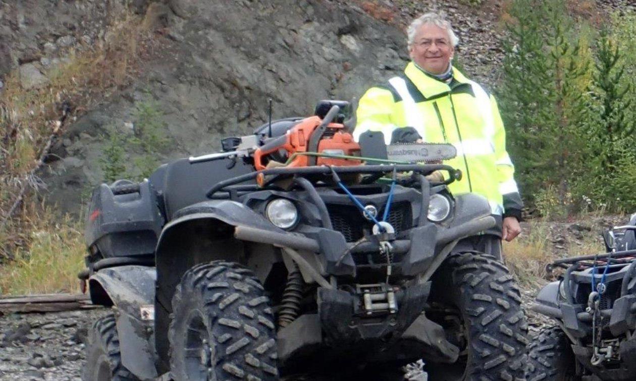 Chris D’Silva stands beside his ATV in front of a mountain while wearing a reflective green jacket. 
