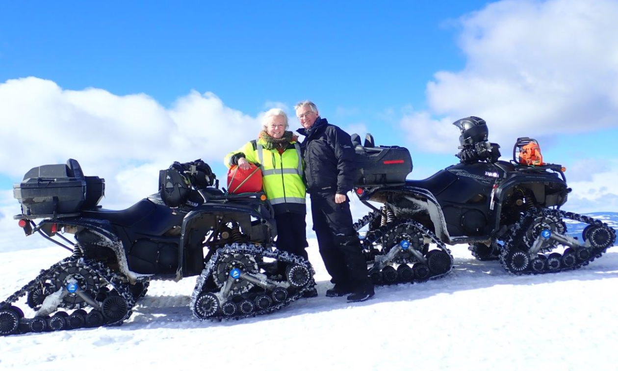 Chris and Jane D’Silva stand next to their ATVs with tracks on the snow beneath blue skies. 
