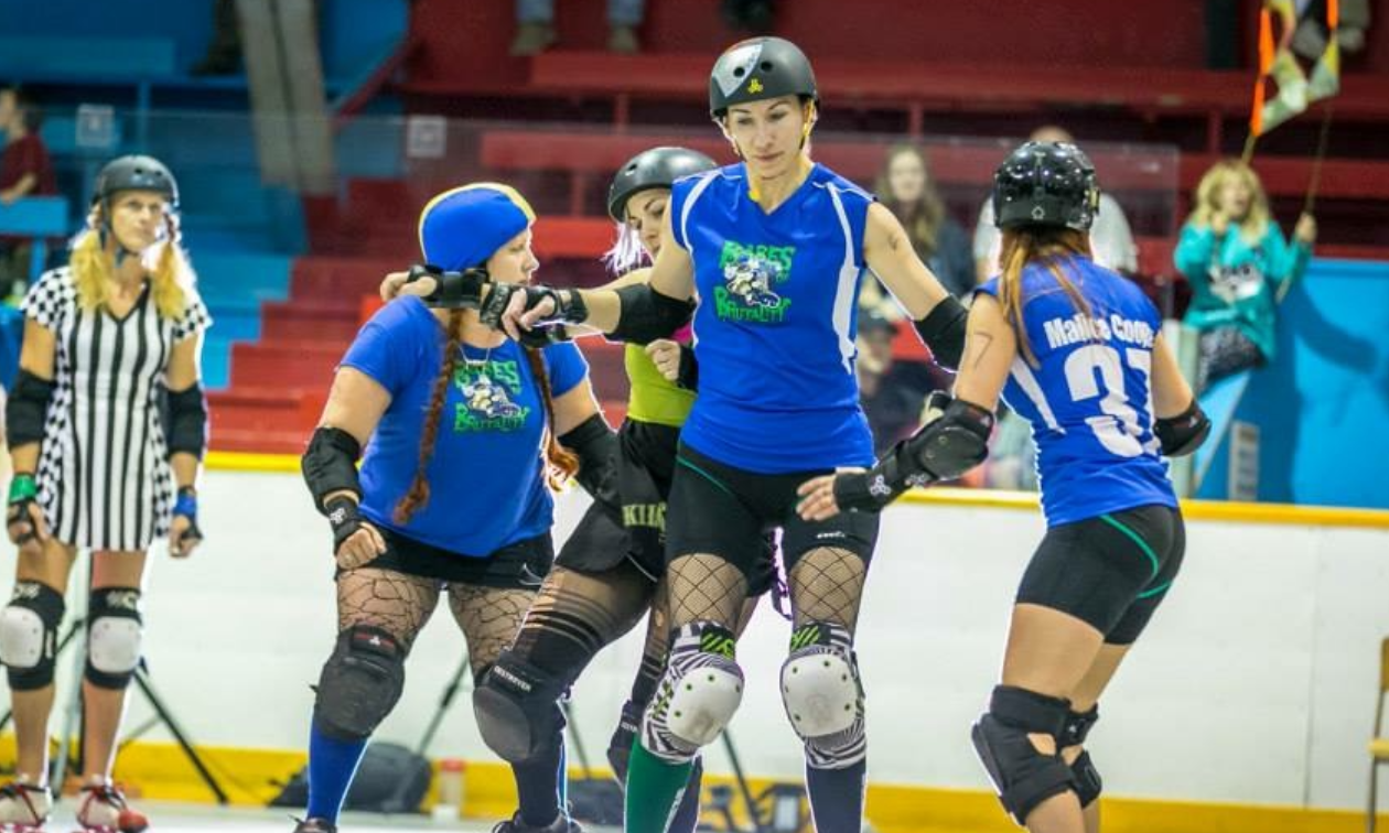 Alysha Vlahovich stands next to teammates on roller skates in an indoor rink while wearing blue. 