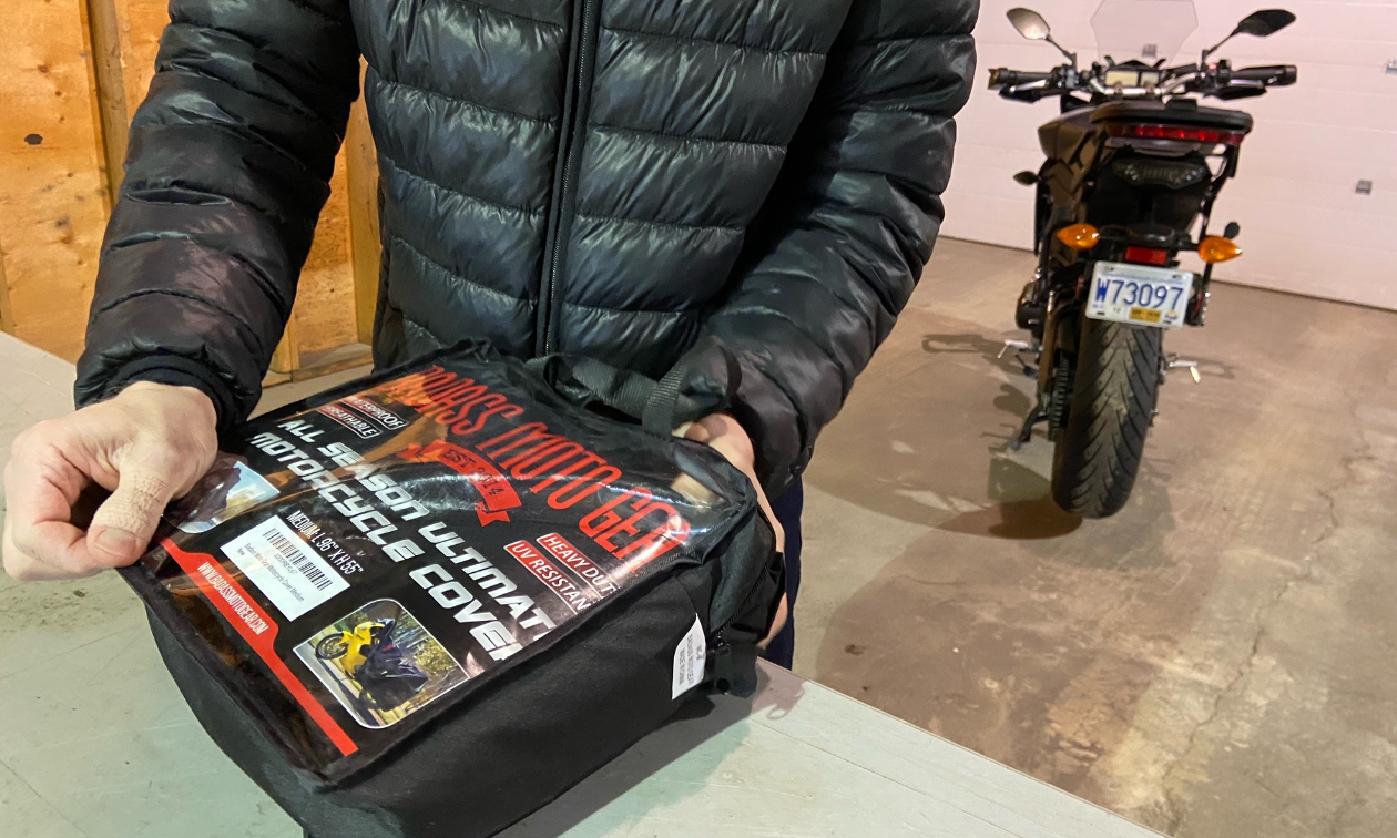 A man in a black jacket opens a package containing a Badass Moto Ultimate All Wx Waterproof Motorcycle Cover while a 2015 Yamaha FJ-09 motorcycle is parked behind him.