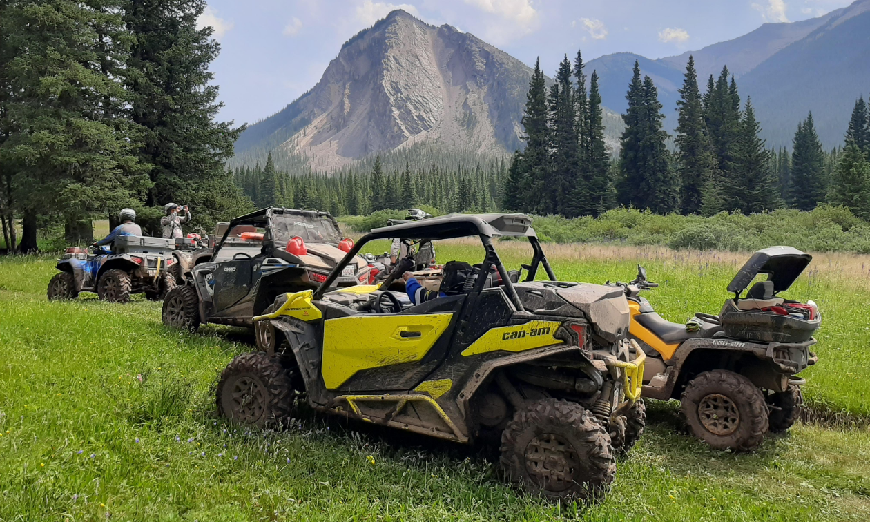 Four side-by-side ATVs are parked in a glade while a mountain juts out of the ground in the distance. 