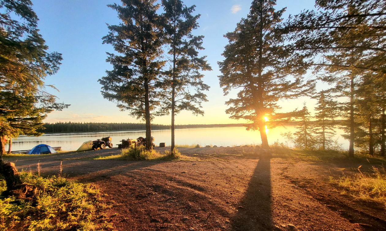Two ATVs are parked near a lake as the sun rises between the trees. 