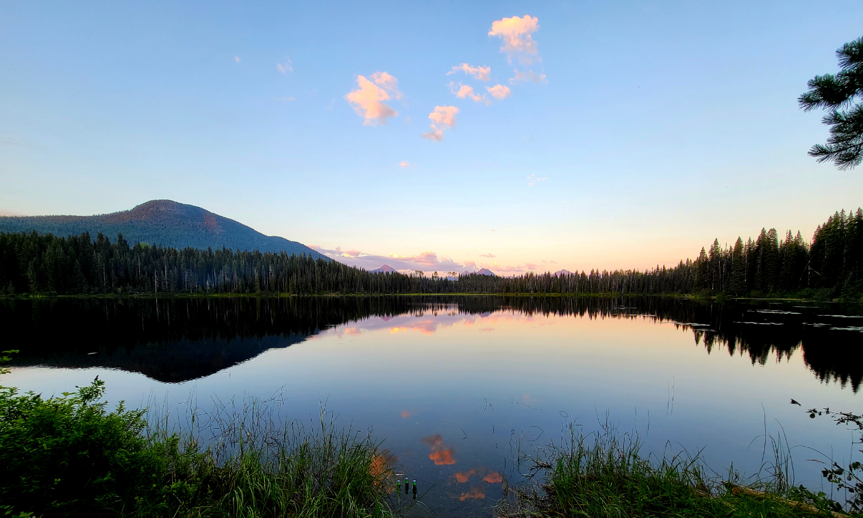 A panorama shot of a still lake with the blue sky and mountains reflected in the water. 