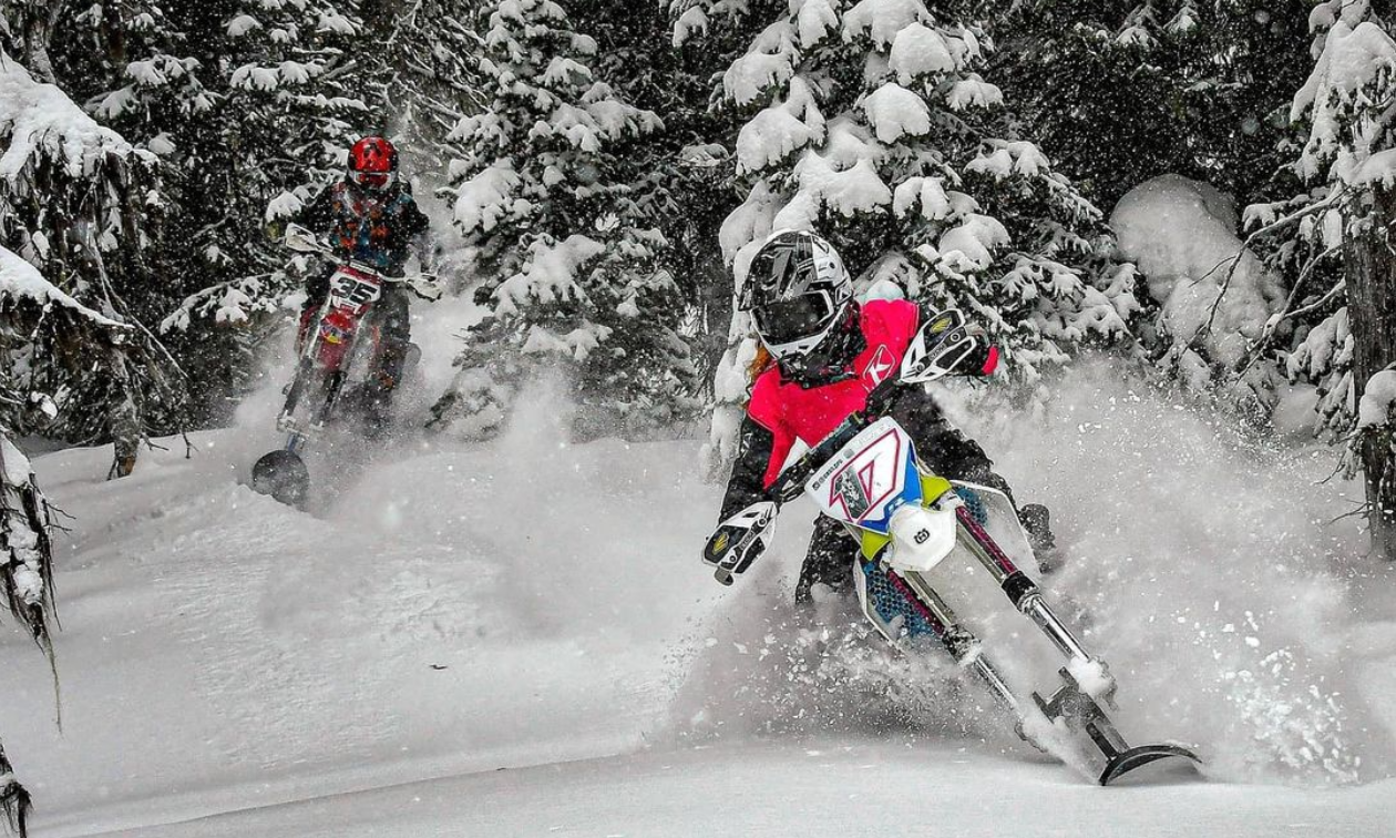 Two snow bike riders scoot through trees in the snow. 