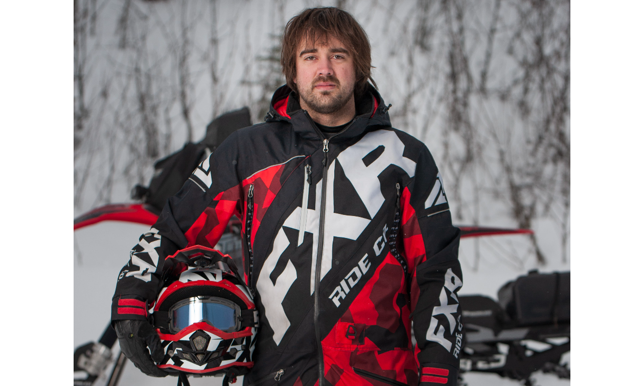 Curtis Hofsink stands straight wearing a red and black FXR jumpsuit and holding his helmet next to his body. 