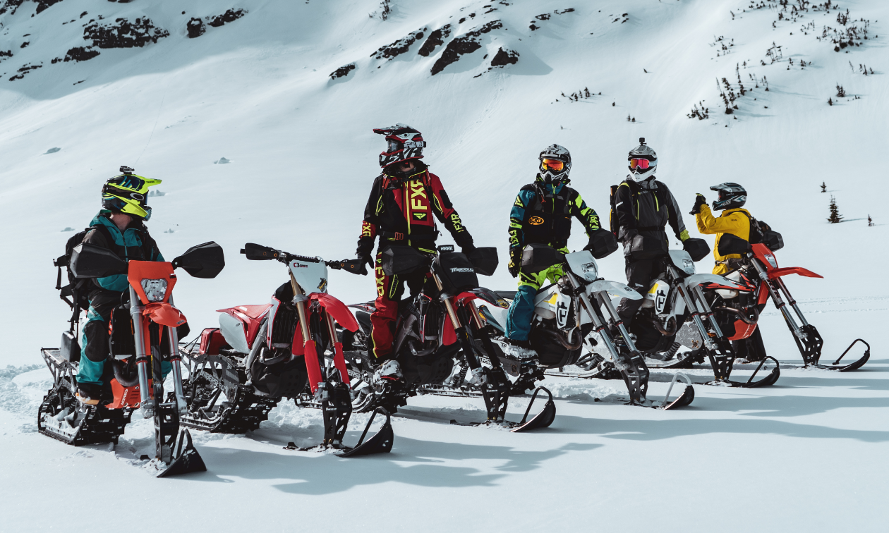 Six snow bikers take a break on top of a snowy mountain. 