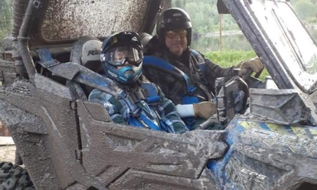 Curtis Riffel and his grandson, Phoenix Mitchell, sit inside a muddy blue side-by-side ATV.