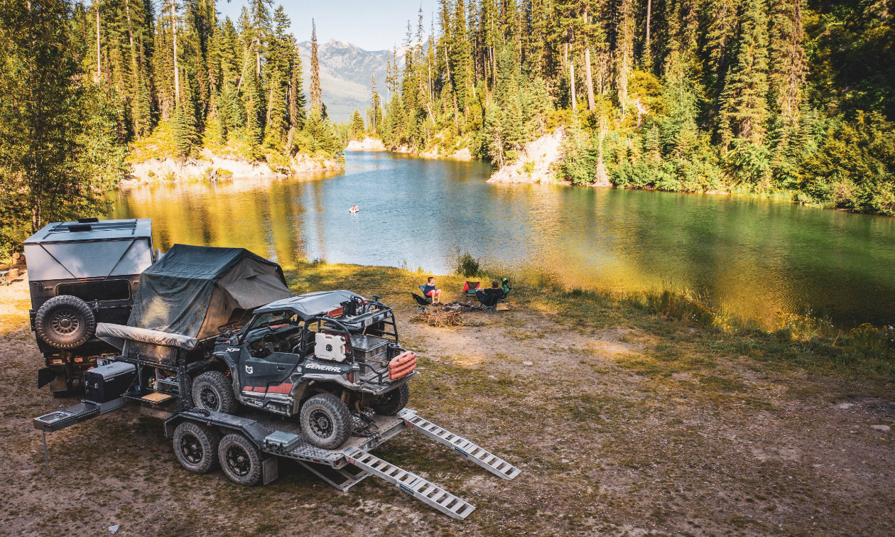 A river in Montana. On the near shoreline, a Dodge Ram 3500, carrying a Patriot Camper TH610 trailer with a Polaris General 1000.