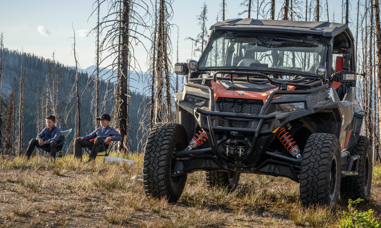 Two boys sit in lawn chairs near some scraggly trees in the background and a Polaris General 1000 in the foreground.