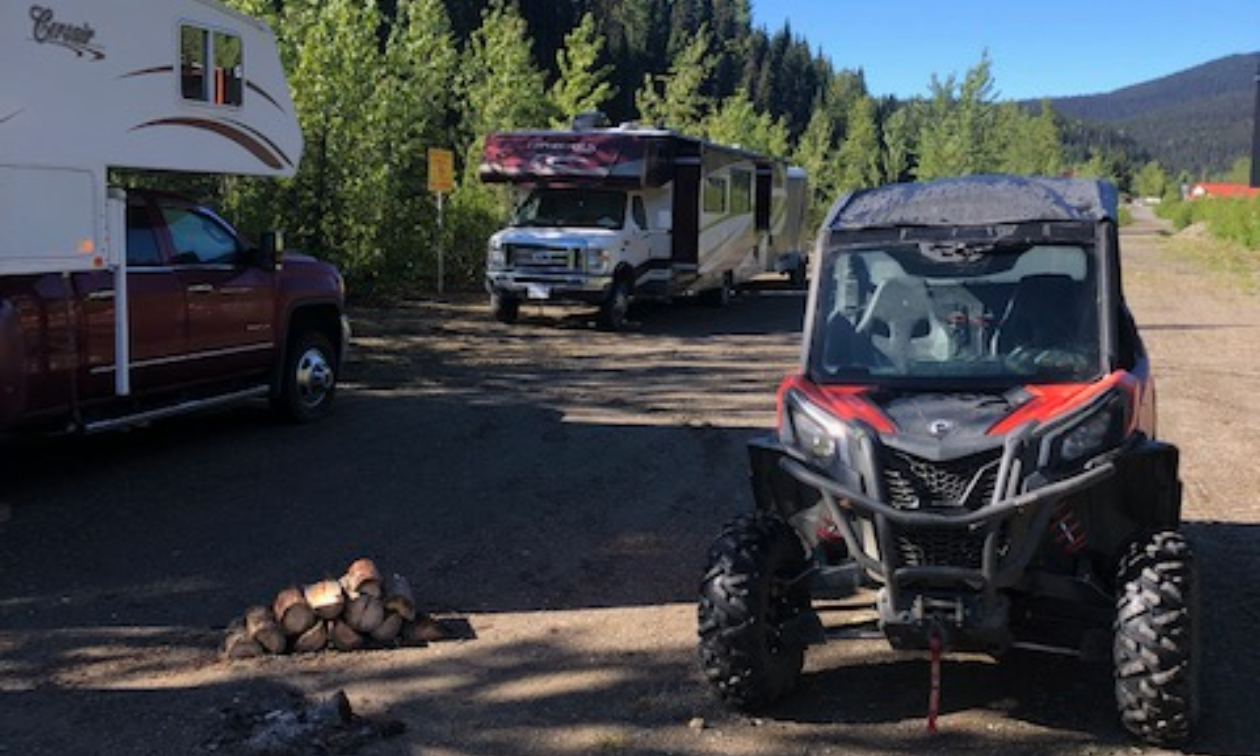 An ATV is parked next to two RVs on a dirt road lined with trees on one side and a field on the other side. 