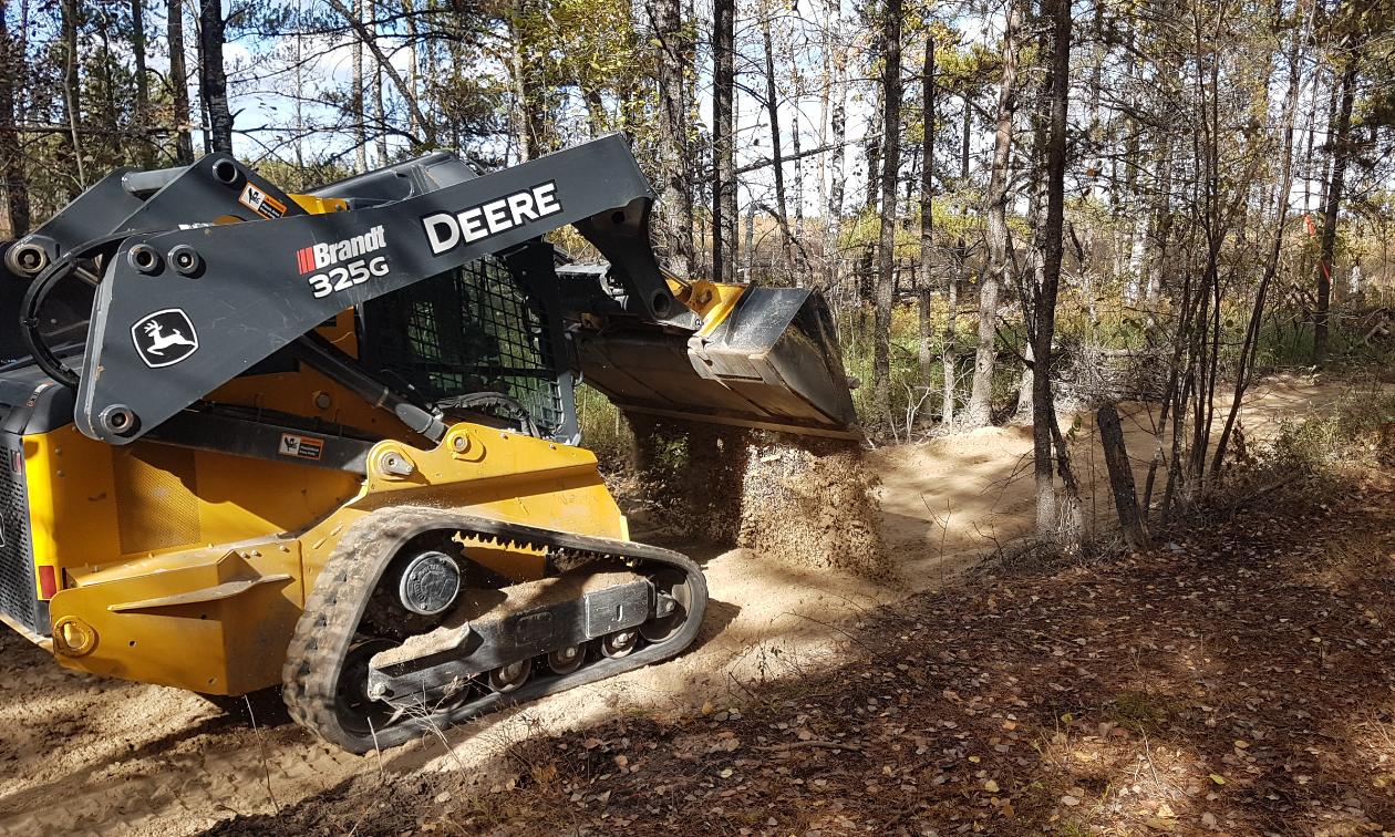A John Deere front loader drops a pile of dirt onto a wet patch of an ATV trail.
