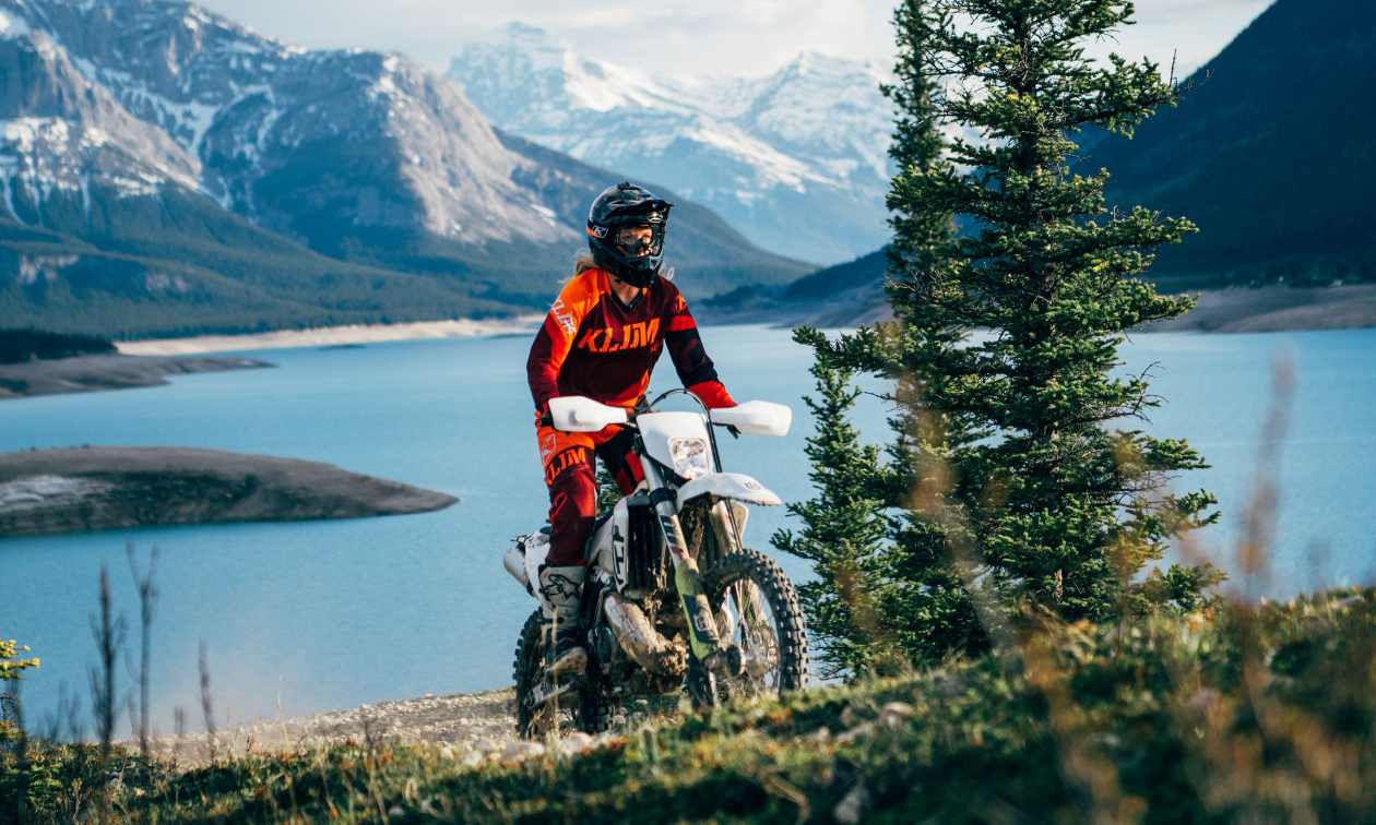 Gloria Cunningham rides a dirt bike up a mountain with a lake and more mountains in the background. 