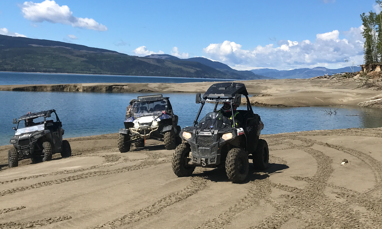 ATVs ride along a sandy shoreline on a sunny day. 