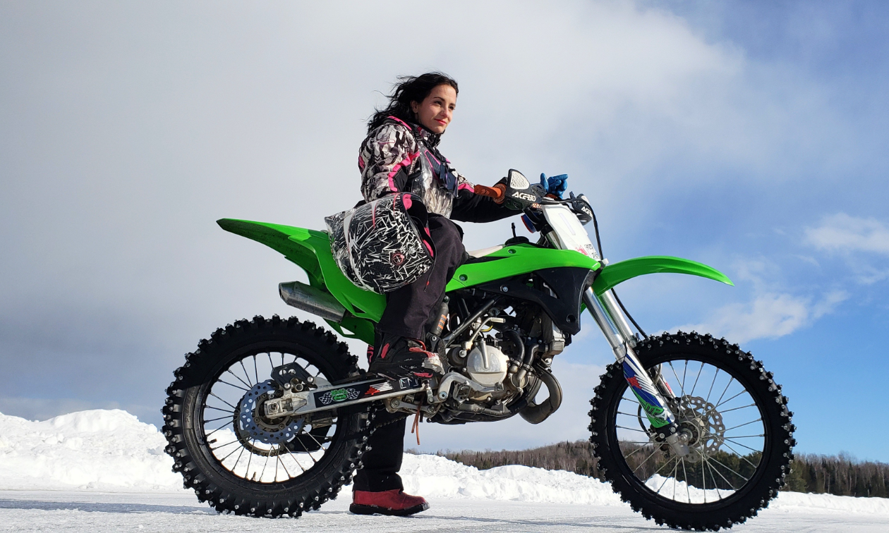 Jessica Rainville poses on a green dirt bike with clouds and mountains in the distance. 
