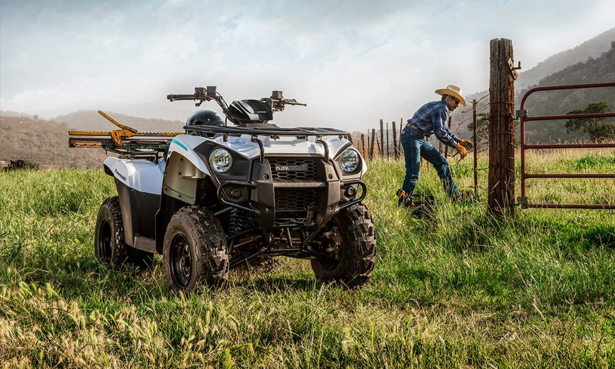 A grey Kawasaki Brute Force ATV in the foreground with a farmer working on a fence in the background.