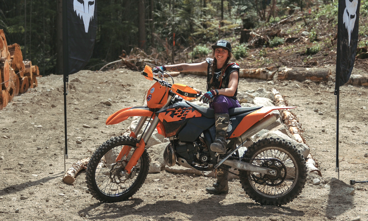 Kirsten Patton smiles on her orange KTM dirt bike in a dirt parking lot next to some Fox flags. 