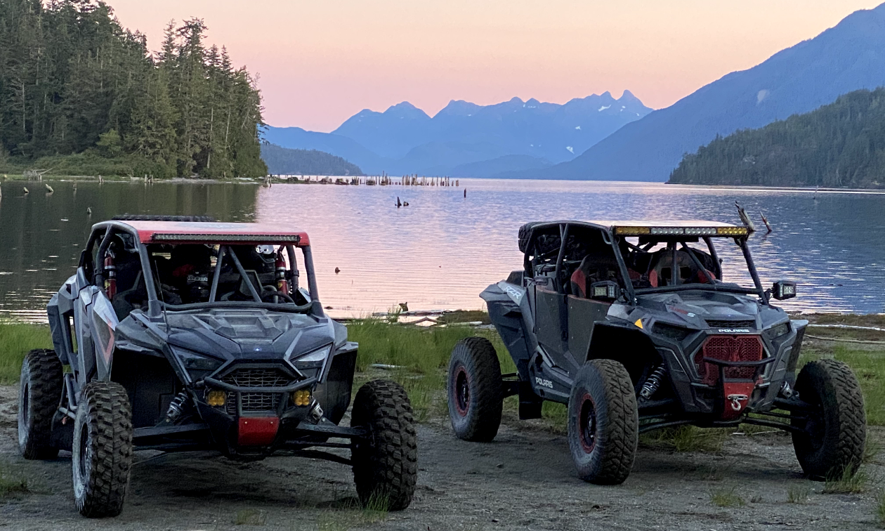 Two side-by-side ATVs next to a lake at dusk with a pink sunset over mountains in the background. 