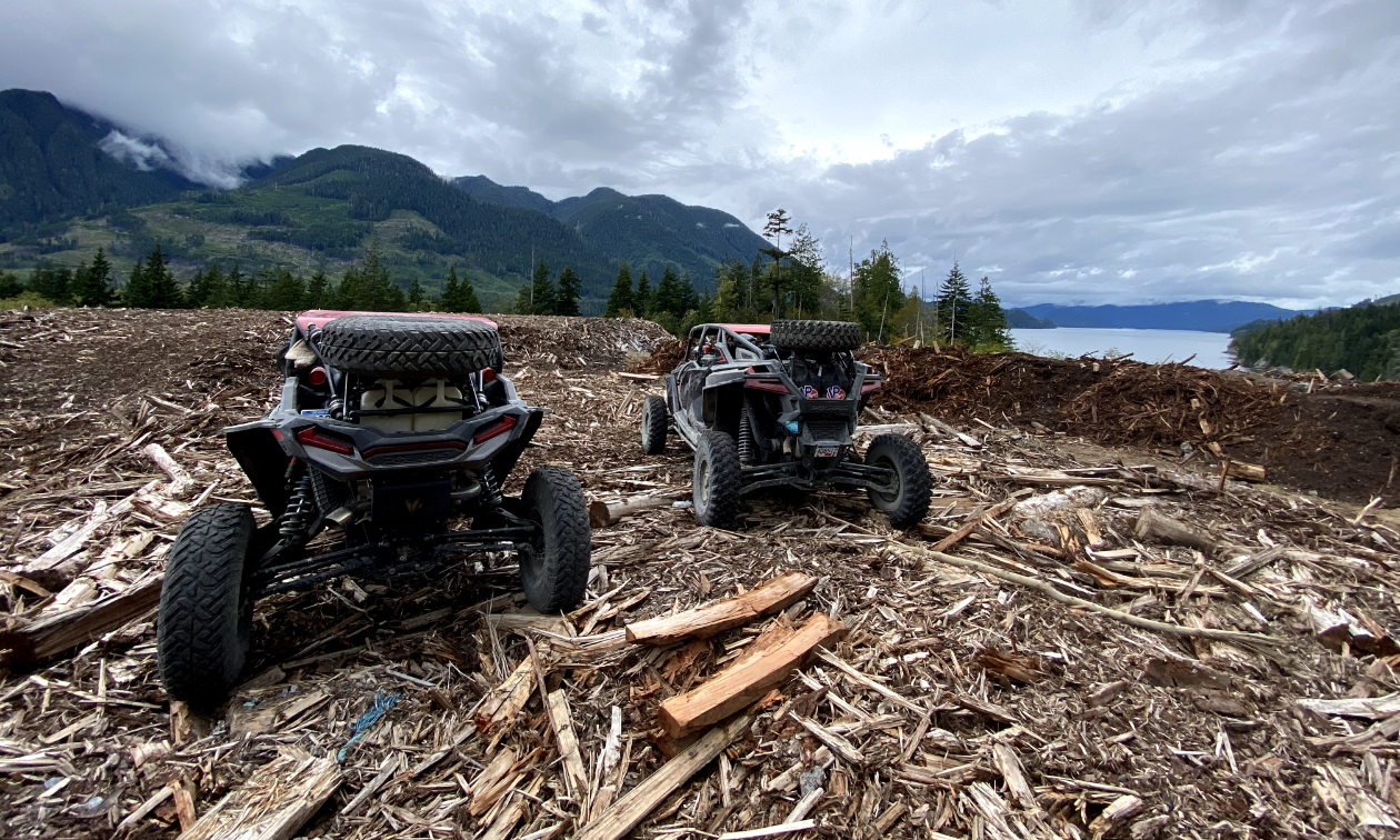 Two ATVs ride over a long expanse of wood chips on a mountain. 