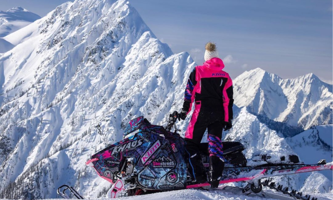 A woman wearing pink and black clothing has her back to the camera as she stands on top of a snow-capped mountain with her pink and black snowmobile with the words “She Shreds” written on the side. 
