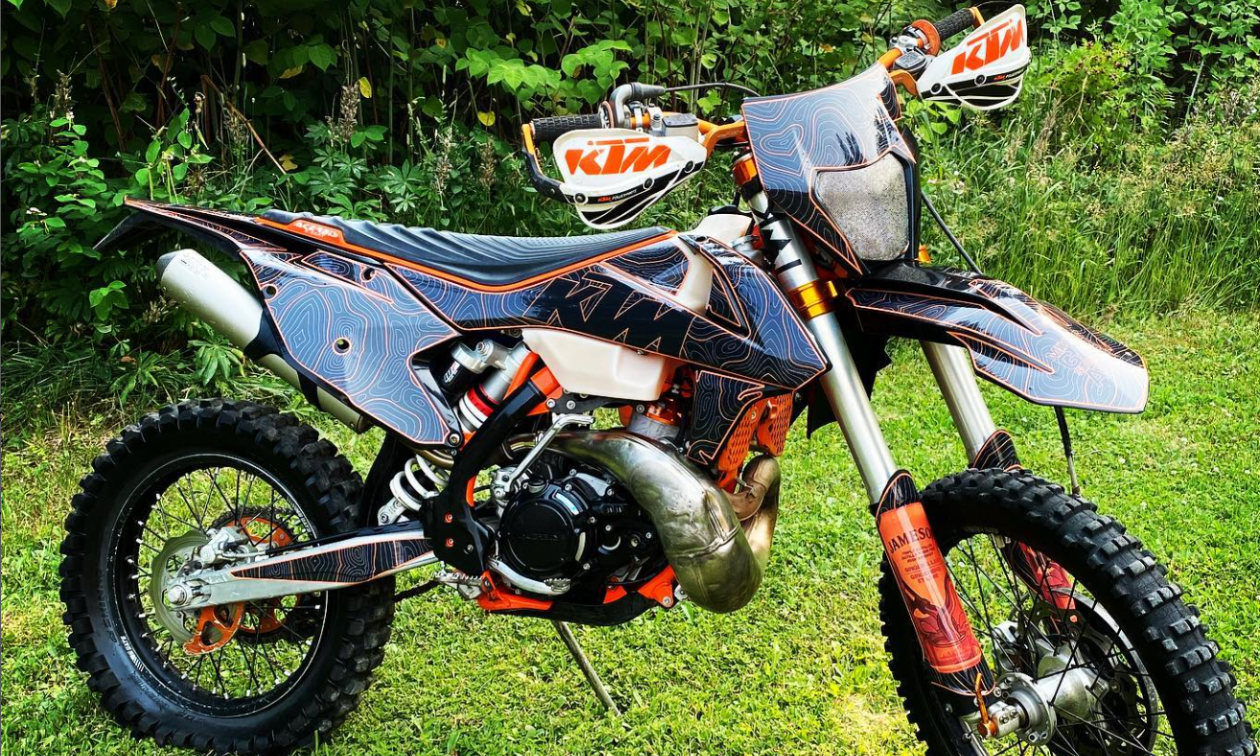 An orange and black wrapped dirt bike shines in the sun on green grass. 
