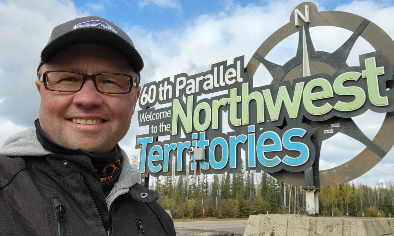Craig Oldfield stands next to a sign that says 60th Parallel - Welcome to the Northwest Territories. 