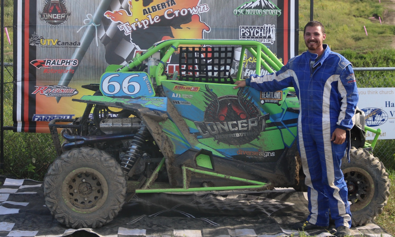 Scott Kreiser stands next to his neon green and blue 2016 Polaris RZR Turbo on a checkered flag tarp in front of a Triple Crown Race Series sign. 