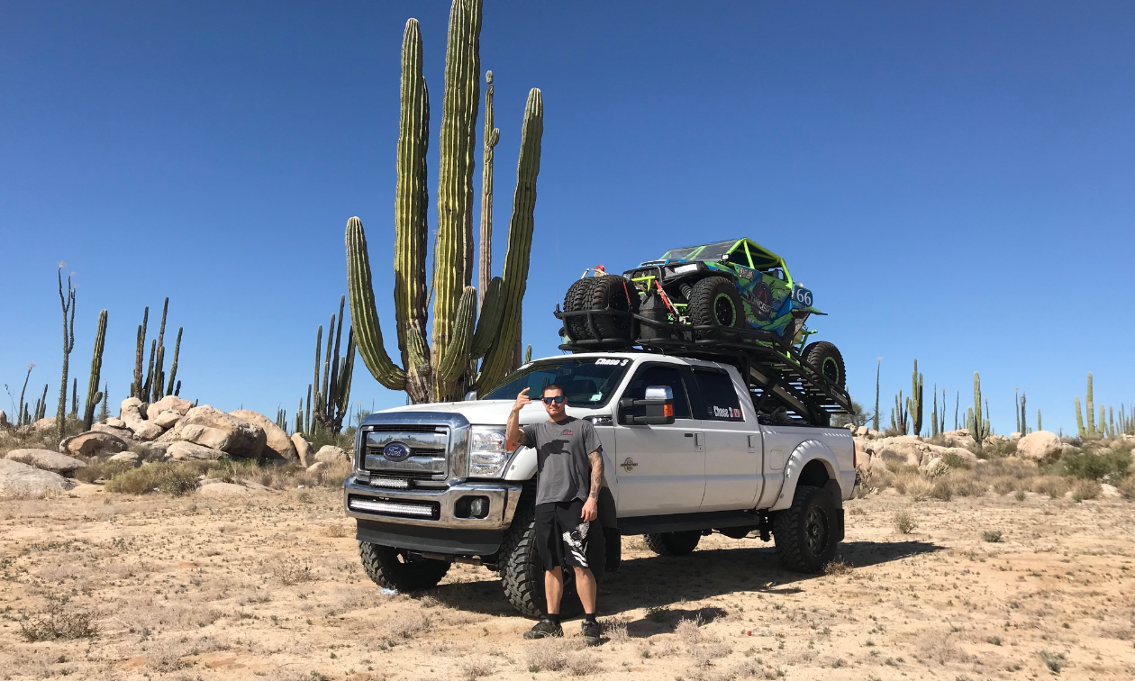 Scott Kreiser stands in front of a white truck with his Polaris RZR loaded into the back. A big cactus is in the desert background. 