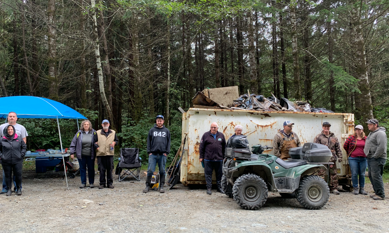 A group of ATVers gather around a dumpster full of garbage in the forest. 