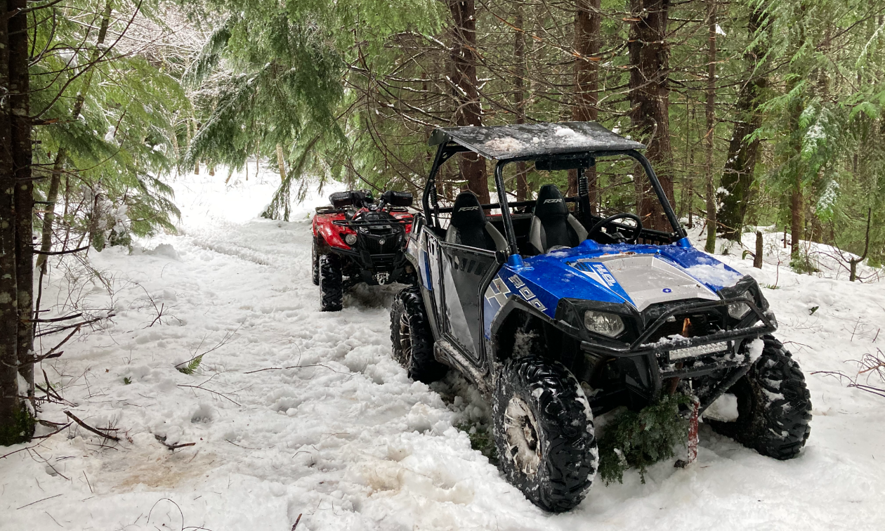 A blue side-by-side ATV is parked on a snowy trail between trees in the forest. 