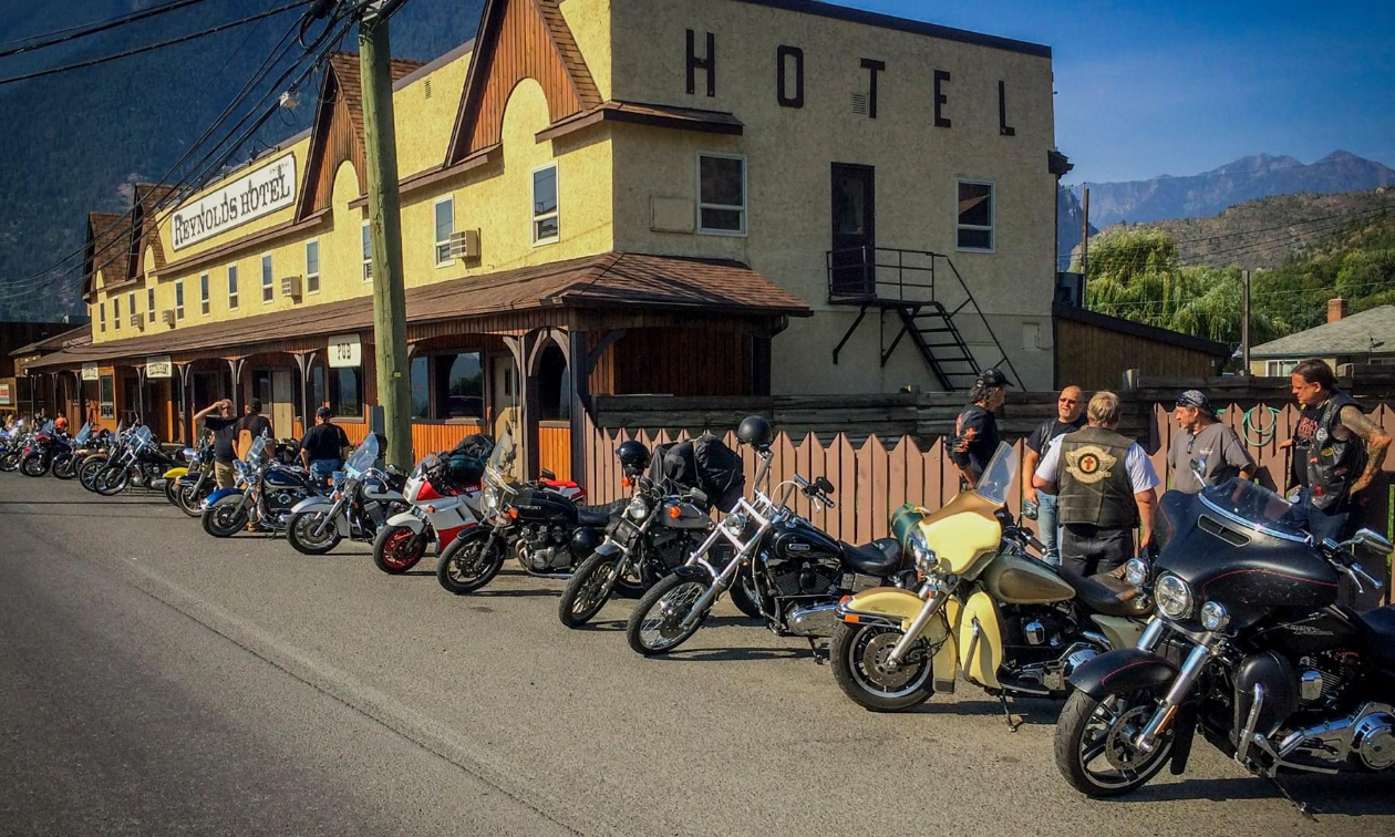 A long row of vintage motorcycles are lined up in front of an old yellow hotel. 