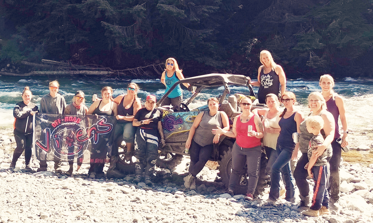 A large group of women stand on a sandy beach in front of and on top of an ATV side-by-side. Chipmunk River flows in the background.