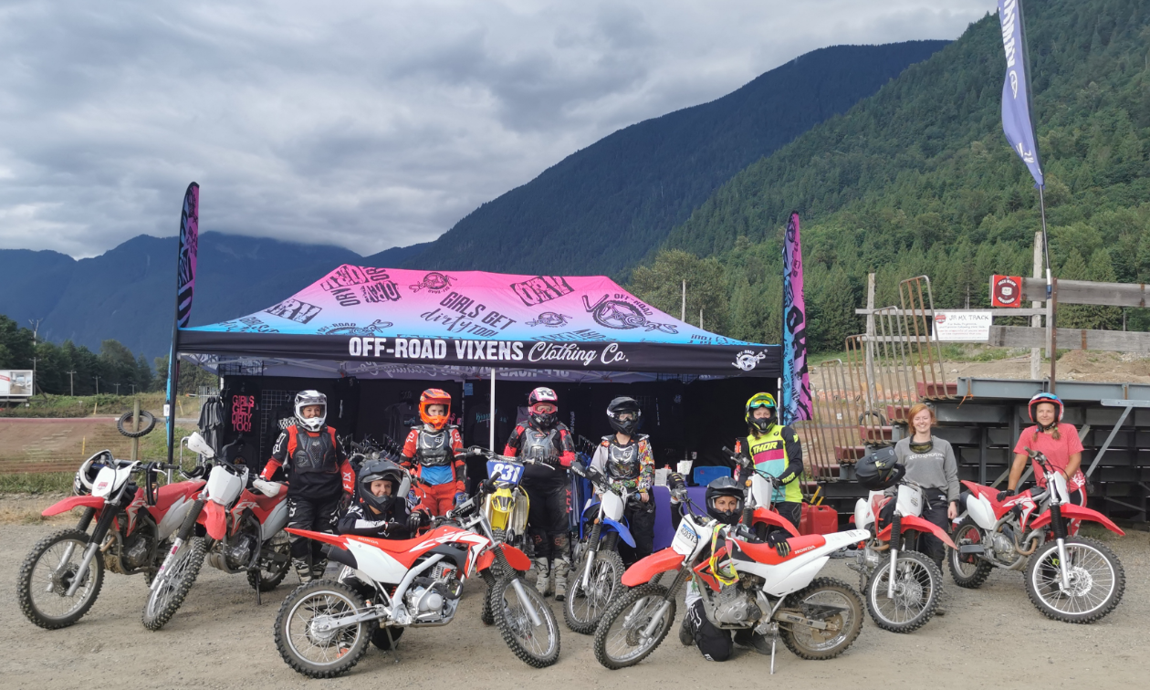 A group of female motocross riders pose in front of a Vixens Canada booth next to their dirt bikes. 