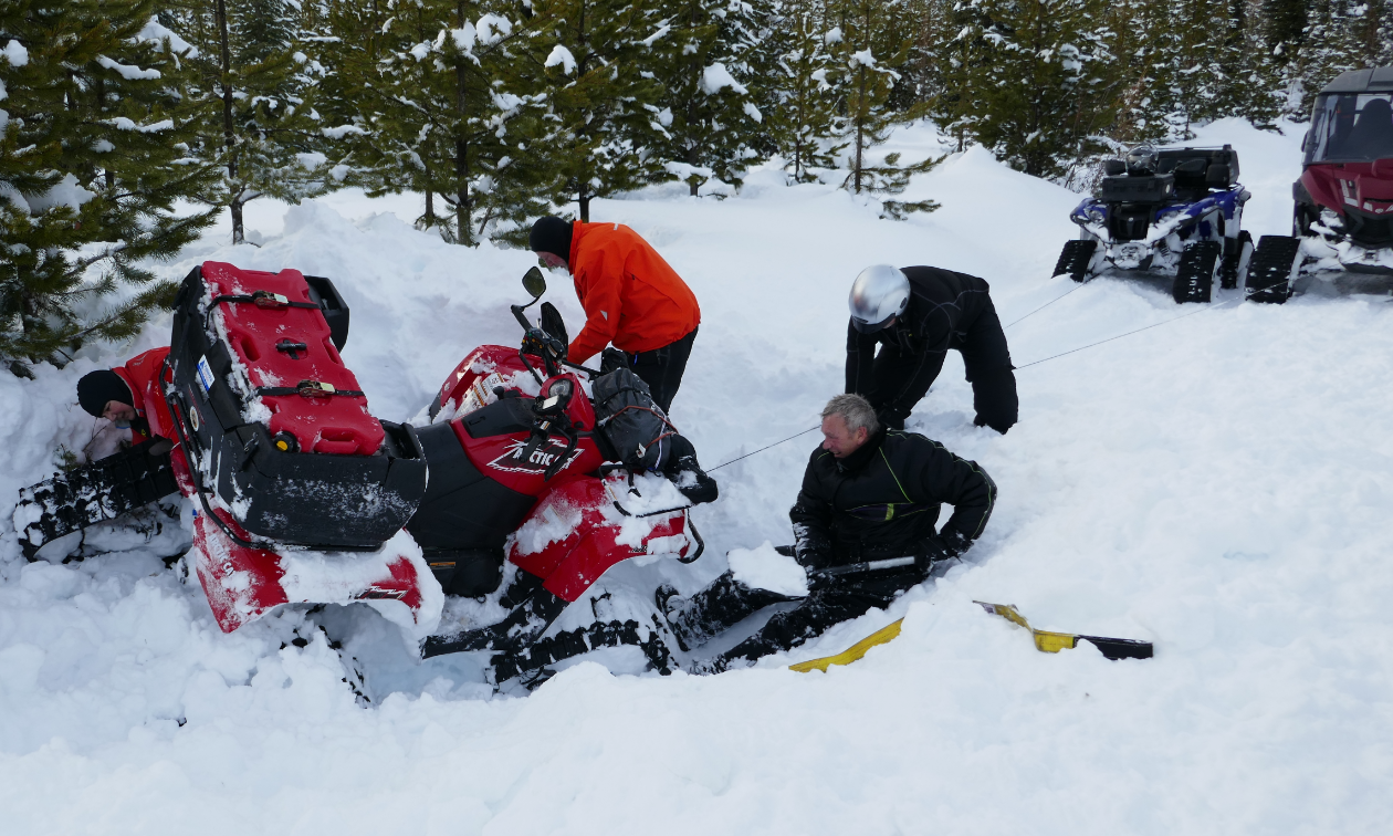 Several snowmobilers hook a winch to an ATV that is stuck in deep snow. 
