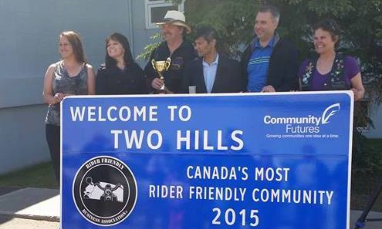 A group of people pose in front of a big blue sign that says Welcome to Two Hills, Canada’s most rider friendly community 2015. 
