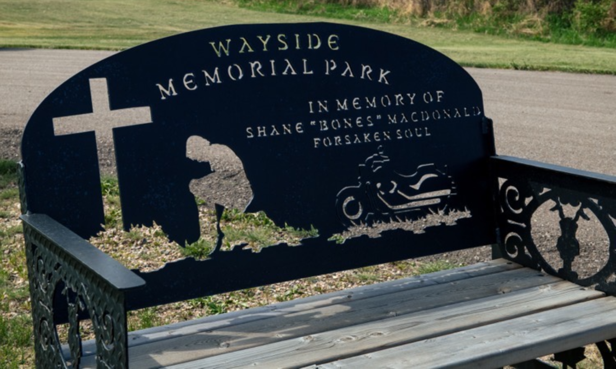 A black park bench that shows a motorcyclist kneeling next to a cross and says Wayside Memorial Park. 