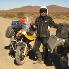 Photo of a guy in a black outfit and white helmet standing in the desert beside a black motorcycle and a yellow motorcycle.