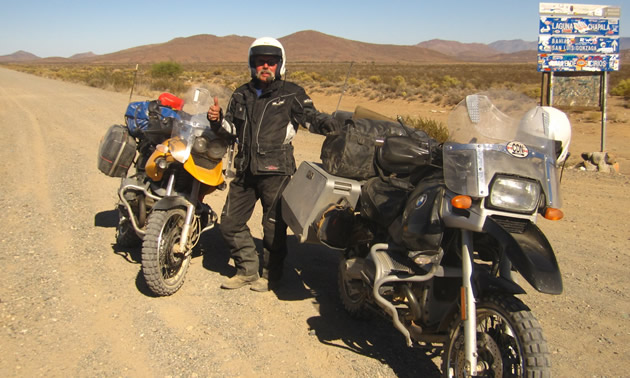 Photo of a guy in a black outfit and white helmet standing in the desert beside a black motorcycle and a yellow motorcycle.