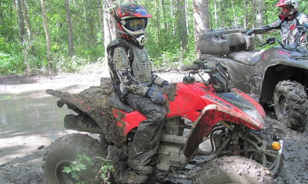 Photo of a young boy sitting on a red quad beside a large mud puddle.