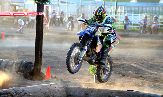 A guy in blue and green riding gear jumping a blue dirt bike over a log on a track. 