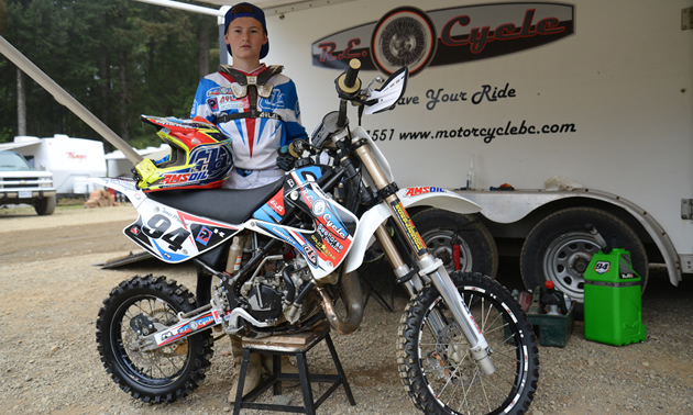A young boy dressed in full moto gear standing by a race dirt bike. 
