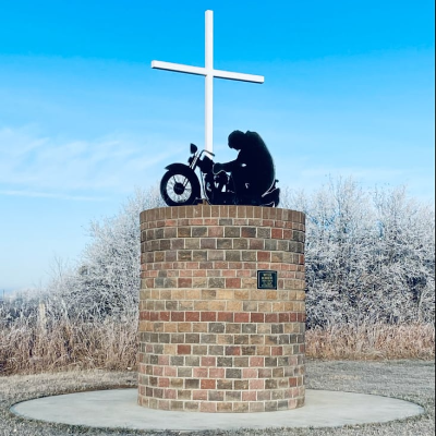 A brick-built circular base showcases a blackened silhouette of a motorcyclist kneeling beside a large white cross. 