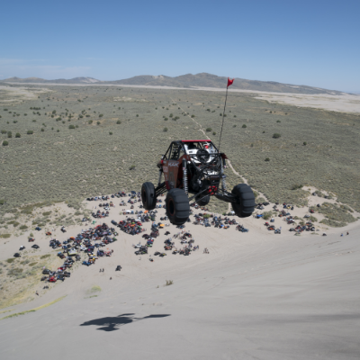 A UTV sails through the air over a steep decline on a sand dune. Many people and vehicles look like ants at the base of the hill.