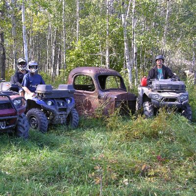 Three quadders line up their vehicles next to a rusted piece of a truck in the woods.