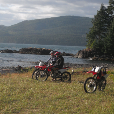 Two dirt bikers sit on their bikes and take in the scenery of ocean, mountains and trees. 