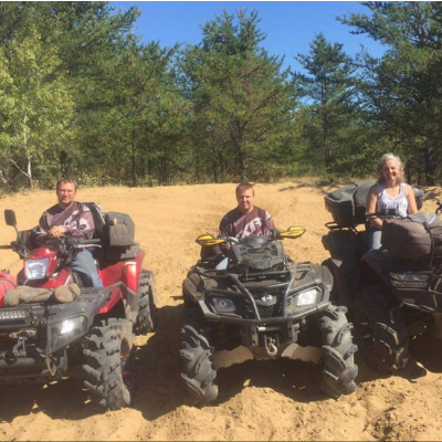(L to R) Darcy, Ryan and Bernice Ruf are sitting on their three respective quads on a dirt patch with green trees behind them.