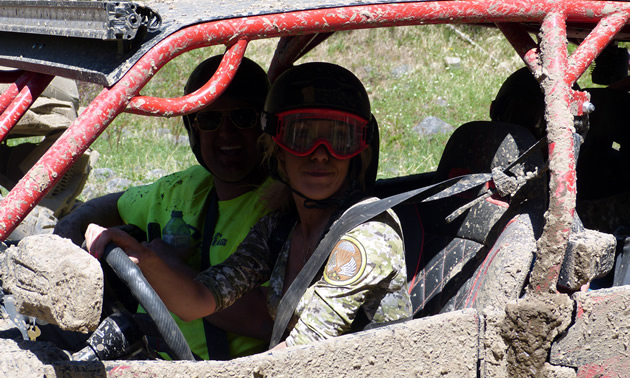 A young woman and man riding in a muddy side by side. 