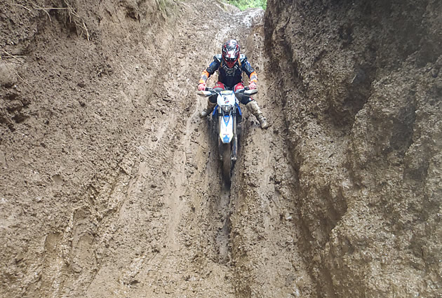 A motorcycle rider going through a deep groove in a muddy bank. 